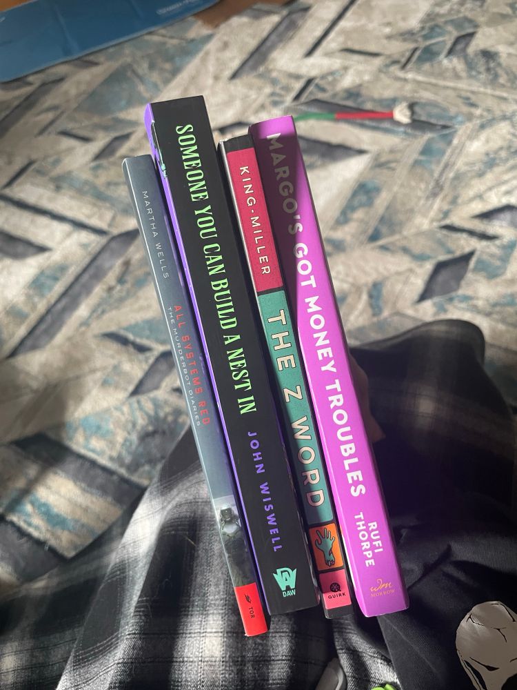 Book spines showing, from left to right: 1. All Systems Red - Martha Wells 2. Someone You Can Build a Nest In - John Wiswell 3. The Z Word - King-Miller 4. Margo’s Got Money Troubles - Rufi Thorpe