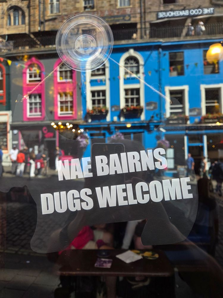 Sign in the window of the Bow Bar, "Nae bairns, dogs welcome". Reflections of buildings opposite.