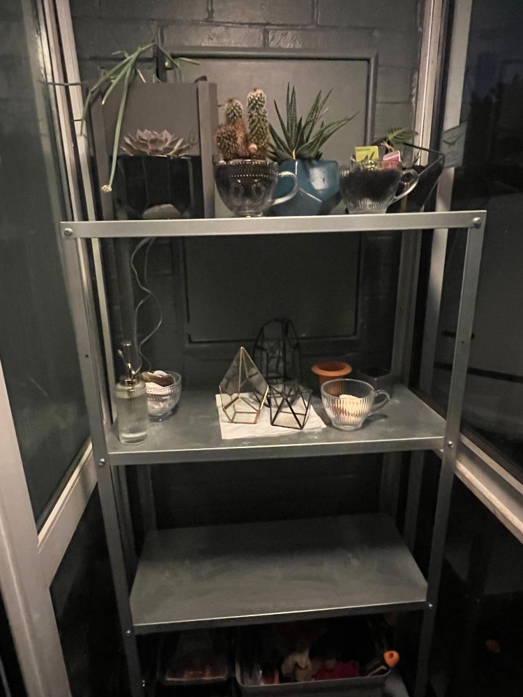 The metal shelf in our front porch, which is mosely glass except for theside walls. The bottom holds gardening tools and propagators. the nect sh lf up is currently empty, the shelf above that has some glass terrariums, containers and cups that are currently empty after my work. and the top shelf has some plants in pots and glass cups. It's dark outside.