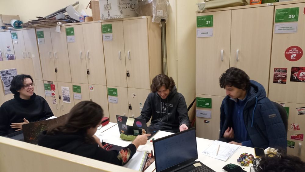 Cinc persones sentades al voltant d'una taula jugant a rol, amb ordinadors, paper i daus a sobre 