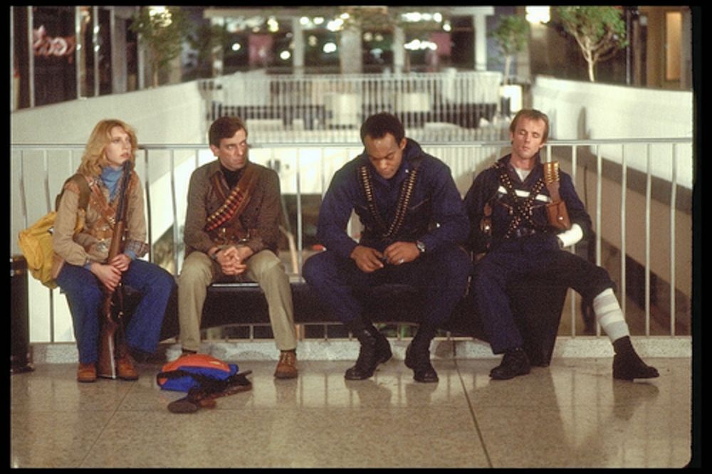 Still from Dawn of the Dead (1978). The four main protagonists (Francine, Steven/"Flyboy", Peter, and Roger) are sitting on a bench in front of railings overlooking a mall courtyard. Francine is holding a rifle and the others are wearing ammo belts around their necks. Roger's forearm and leg are bandaged. They look very tired.