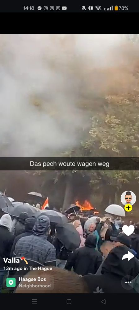 "A crowd of people holding umbrellas gathers outdoors in The Hague, with thick black smoke and flames visible in the background. The text overlay in Dutch reads ‘Das pech woute wagen weg,’ and the location tag indicates ‘Haagse Bos Neighborhood.’