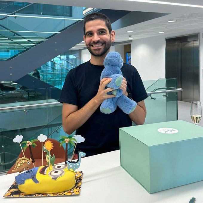 Vicente stands behind a table holding a dinosaur plush toy. In front of him is a table which contains the blue-green box from which the dinosaur plush appeared, and a minions celebration cake which has dinosaur decorations on cocktail sticks on top of it.