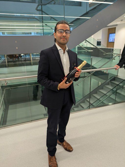Akaash stands in front of some stairs, holding a bottle of champagne whilst wearing a suit.