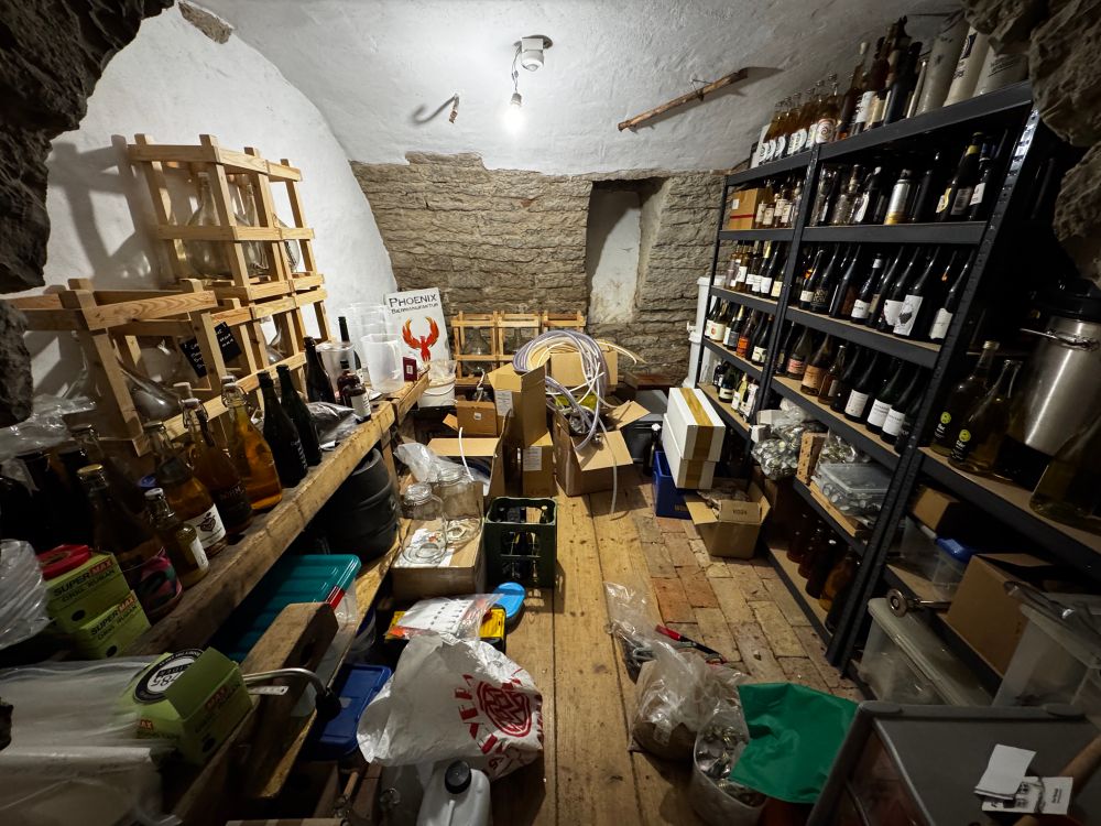 A small vaulted cellar, shelves on the right filled with bottles, an old woodworking bench on the left stacked with glass carboys filled with spirits, and at the back the same. The floor is covered in boxes, crates and sacks. 