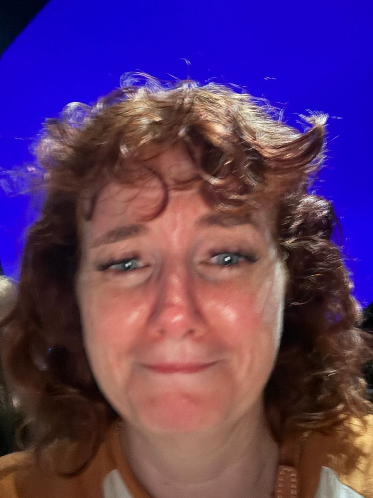 A white woman with tragically curly and short red bangs in front of like a blue screen!?? She looks way sadder about it than this actually merits