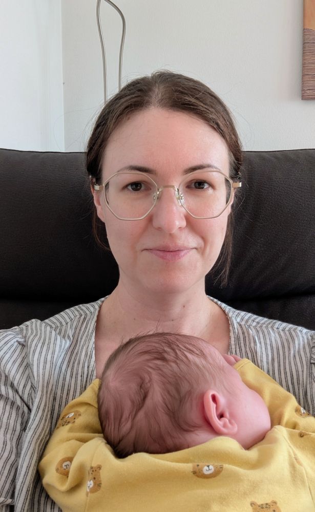 The poster looking at the camera while a one month old baby in a onesie sleeps on her chest.