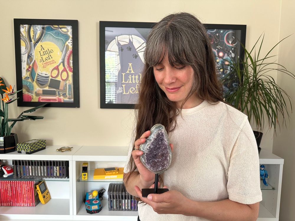 Annie holding and looking at an amethyst rock gifted by popular streamer PapaPupp, in front of three A Little to the Left posters and a display of video game trinkets