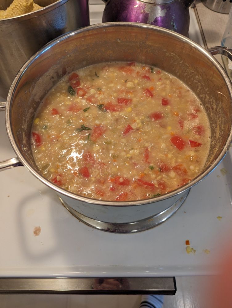 A steel pot full of soup. Corn kernels, chunks of tomato and bits of basil leaves are visible.