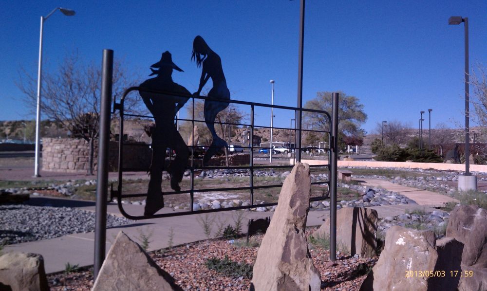 Metal art piece looks like a livestock gate with two silhouettes against it. One a cowboy standing at the gate, and the other a mermaid sitting on the top of the gate.