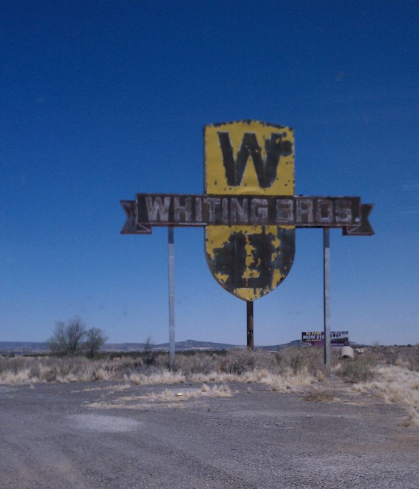 Well worn sign for Whiting Bros. service station against a blue sky. Stands in a foreground of more gravel and weeds than pavement.