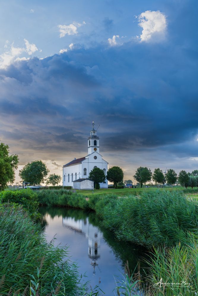 A little white church reflected in the water with a beautiful raincloud over it