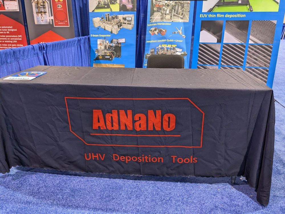 A draped table at a convention, with the table cloth reading AdNaNo -- the joke being it can be read as "Ad? Na, No" -- a question followed by a dismissal. Context around the image shows the booth is actually for UV thin film deposition tools.