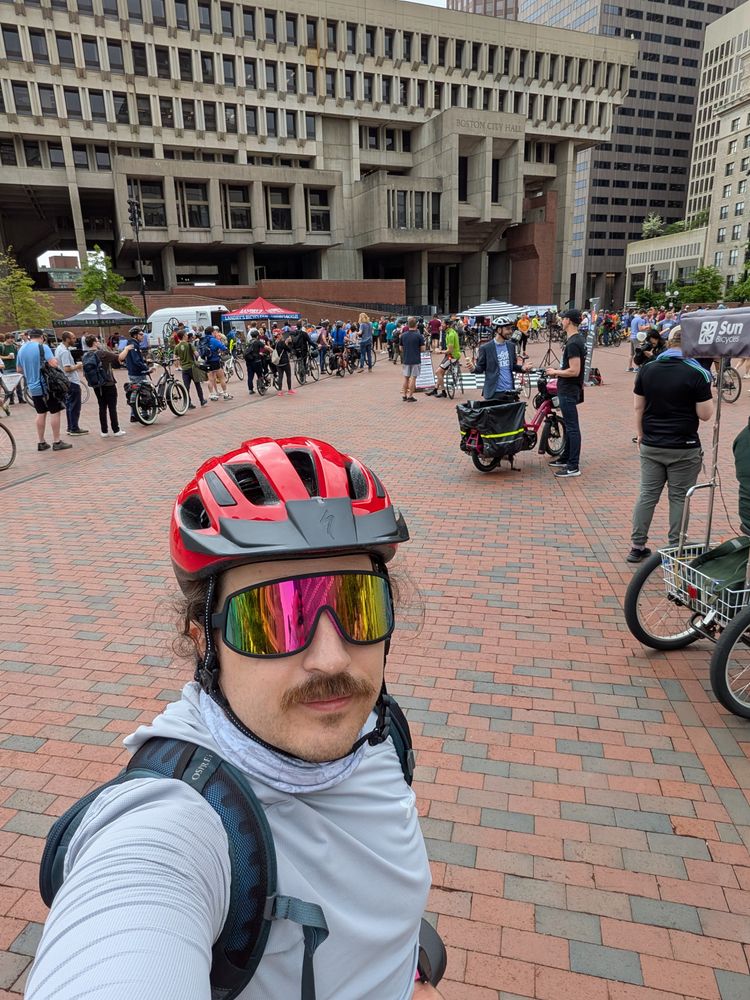 Moustachioed man with a red bike helmet and sunglasses standing in front of Boston City Hall among a crowd of people and bikes.