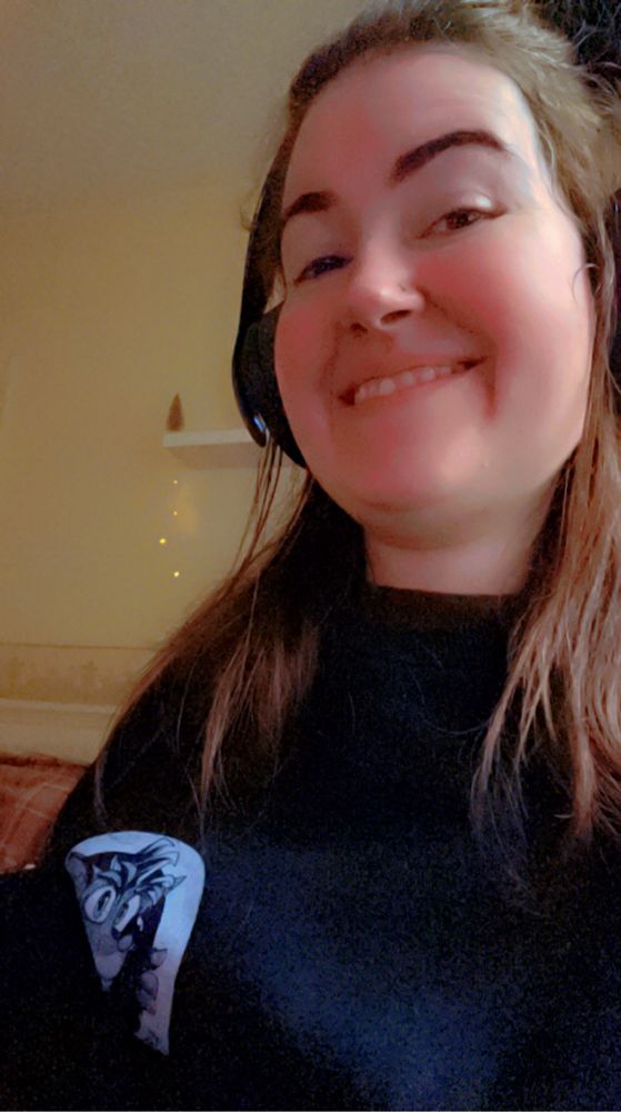 Danielle is wearing a black sweater with a white Crash Bandicoot logo. She has brown hair which is hanging over her shoulders. She has black headphones on and is smiling at the camera 
