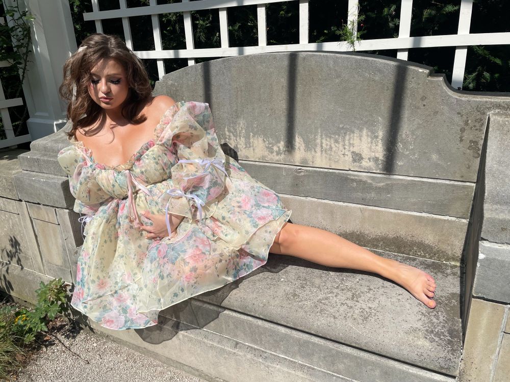 Me in a fancy floral dress on a stone bench outside, caressing my baby bump