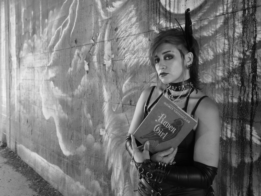 Black and white photo of a woman holding Audrey Niffenegger's book "Raven Girl." The model, dressed in black, has two feathers in her hair, and is standing in front of a mural by Justin Suarez.