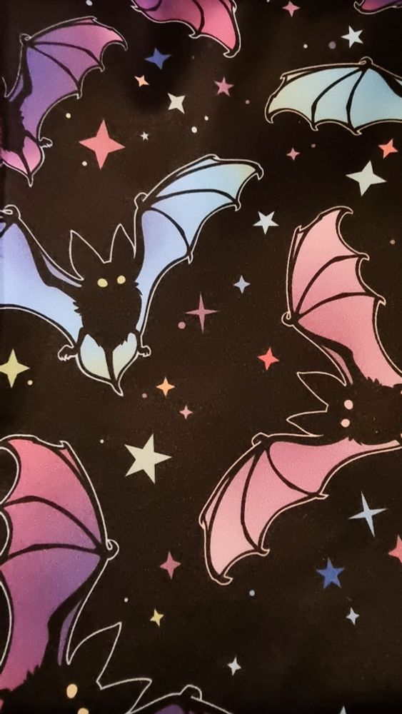 A close up images of some stretch fabric with bats on it. The background of the fabric is black with stars in various colours. The bats have black bodies. Some have wings in shades of blue and teal with yellow eyes and some have wings in shades of purple and pink with pink eyes. 