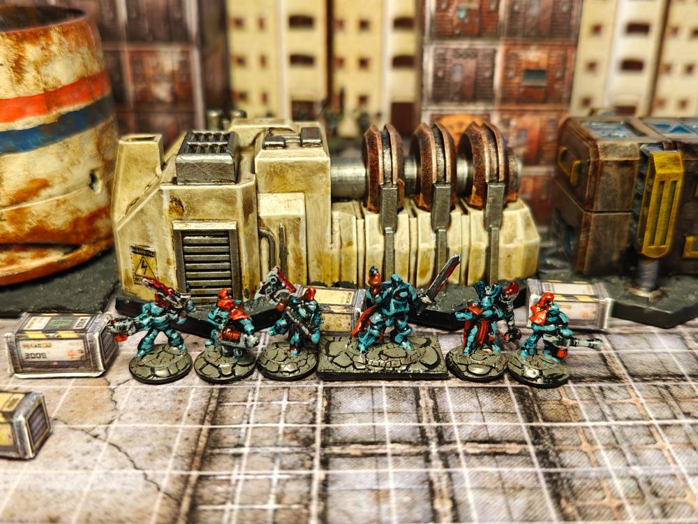 6 painted miniatures in 6mm scale. These represent 6 humanoid creatures in power armour. Their armour is painted turquoise with red accents and they wield a variety of weapons, both melee (axes, swords) and ranged (pistols, machine-guns). The central model is slightly bigger than the others, representing a leader or hero. The miniatures are photographed in front of 6mm scale terrain representing a generator in a sci-fi setting.