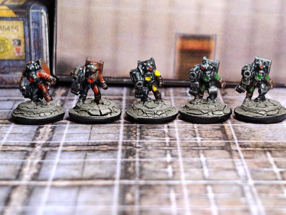 5 painted 6mm-scale miniatures (front view). The miniatures represent robots or cyborgs, they are painted mainly in metallic colours with red, yellow, or green clothes. The two miniatures on the left have rust over the metal parts. The miniatures are photographed over a background of paper terrain