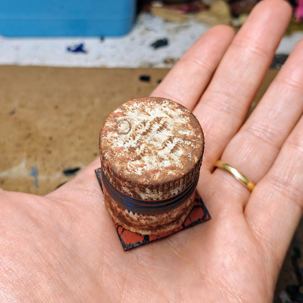 a 6mm scale piece of terrain (view from the top). It has been built gluing together two oil caps. They have been painted in a beige tone with a blue and red line in the middle, then covered in rust. The resulting piece of terrain has been placed on a 3.2cm square base.