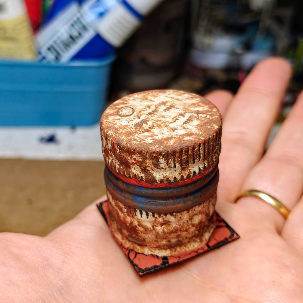 a 6mm scale piece of terrain. It has been built gluing together two oil caps. They have been painted in a beige tone with a blue and red line in the middle, then covered in rust. The resulting piece of terrain has been placed on a 3.2cm square base.