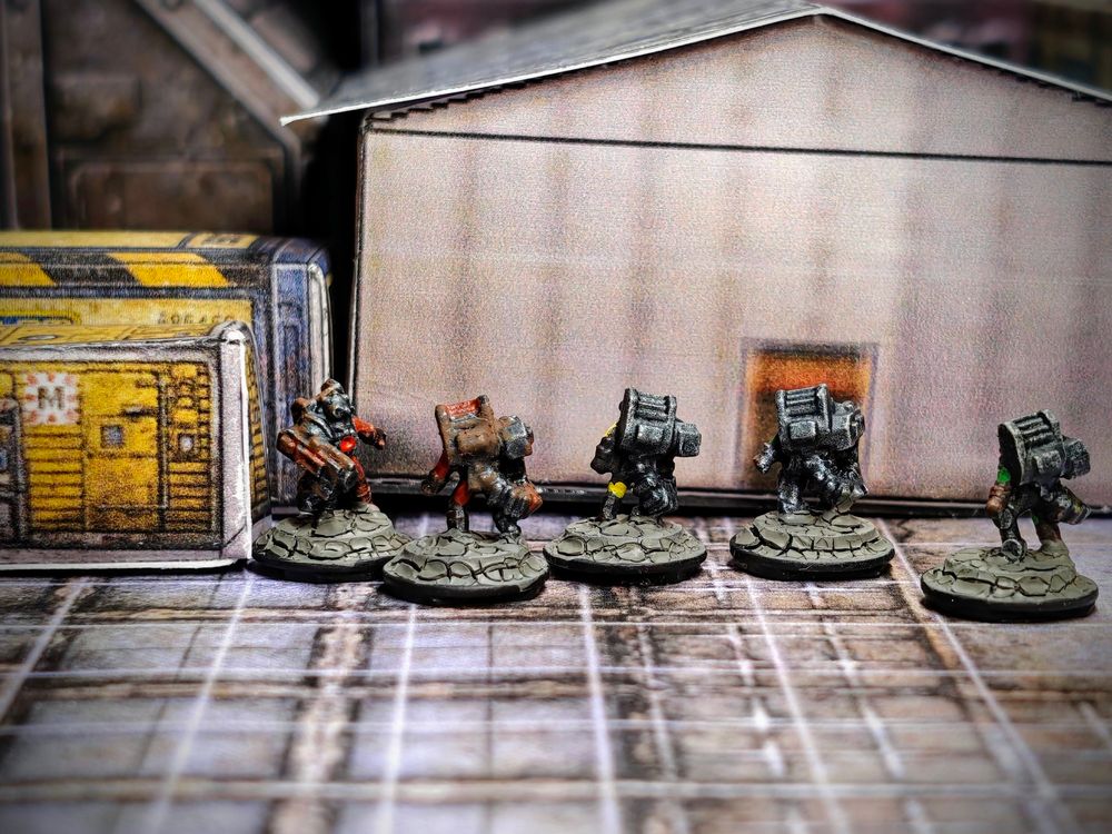 5 painted 6mm-scale miniatures (rear view). The miniatures represent robots or cyborgs, they are painted mainly in metallic colours with red, yellow, or green clothes. The two miniatures on the left have rust over the metal parts. The miniatures are photographed over a background of paper terrain 
