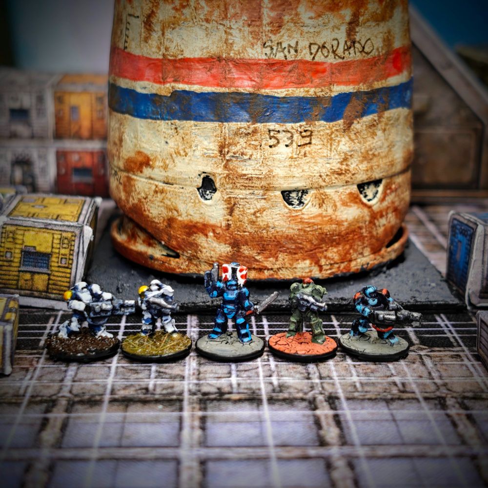 a picture of five painted miniatures in 6mm scale. Left to right, the first two miniatures have a white power armour with a single yellow pauldron; the central miniature has a blue power armour, a white and red back banner, and carries sword & pistol; the fourth miniature has green power armour and a red helmet; the fifth miniature has a turquoise power armour with red pauldrons. They are posed in front of a large (in scale) rusted vent with the name "Sand Dorado" written on it.