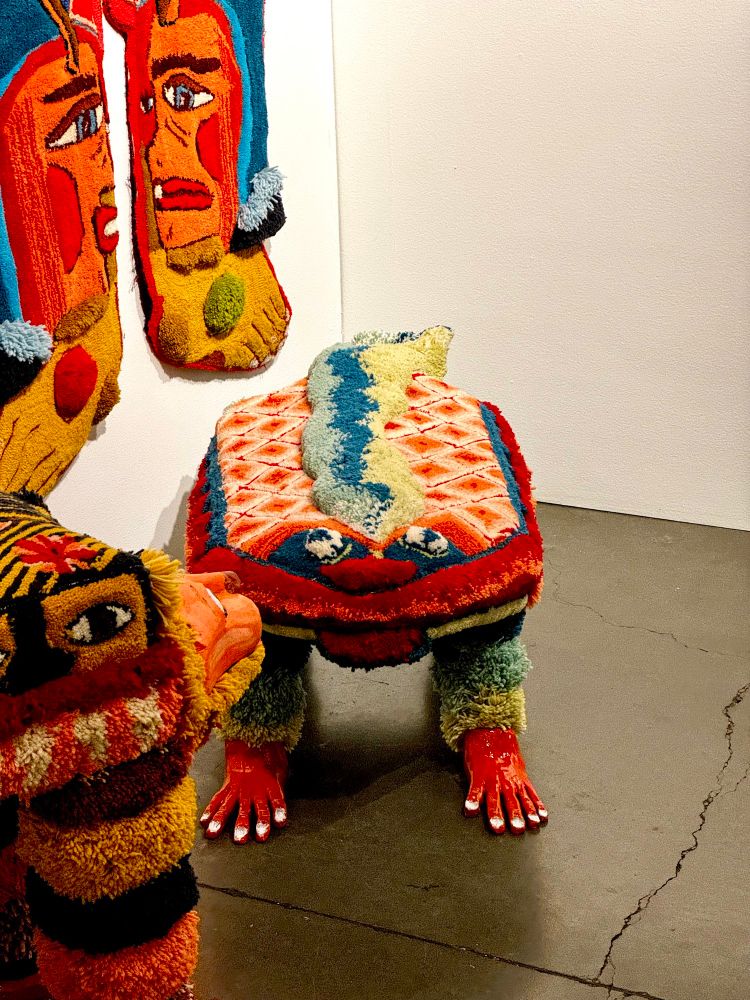 A patterned and colorful shaggy ottoman in the shape of an unknown creature with four legs and a  neckless face with a wide smile. Other colorful carpet creatures surround. 