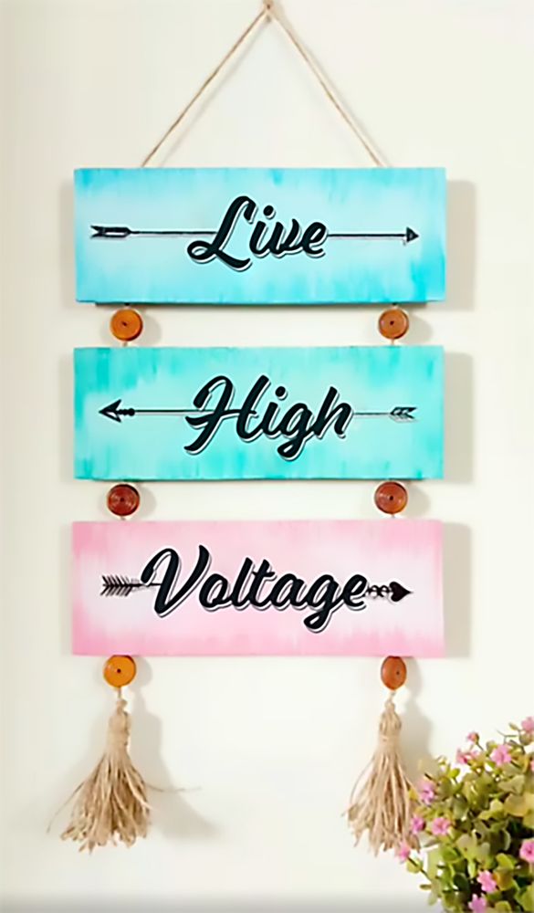 A photo of a colourful affirmative wall hanging in cyan green and pink with macrame styling and photographed in light. The three boards read LIVE HIGH VOLTAGE.