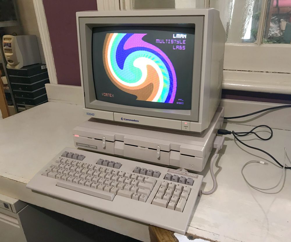 A picture of a Commodore 128DD computer from the mid 1980s. It's a pizzabox style with a separate keyboard and two built-in 5.25" floppy drives. Sitting on top is a Commodore 1084D monitor, displaying a colourful music demo.