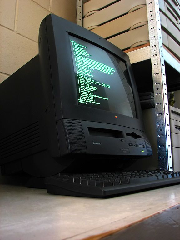 A photo of a black Power Macintosh 5500in a low-lit room, running linux, and showing a shell session with some options listen onscreen. It's a black all-in-one machine with a 15 inch shadow-mask crt, and floppy & CDROM drives on the front, with some volume and brightness controls.
