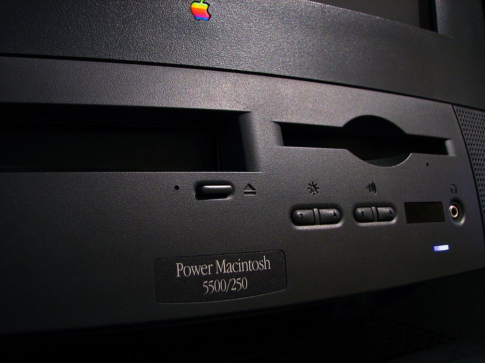 A close-up photo of the front of the black power mac, with rainbow apple logo up top just under the display, a black cdrom tray hole, a floppy disk slot, and some other buttons and jacks. At the bottom is a badge with "Power Macintosh 5500/250" in light garamond text against a black background.