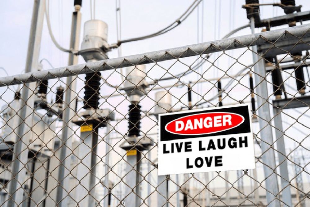 A photo looking up through a chainlink fence at piles and transmission equipment in an electrical substation. There is a sign on the fence stating DANGER LIVE LAUGH LOVE