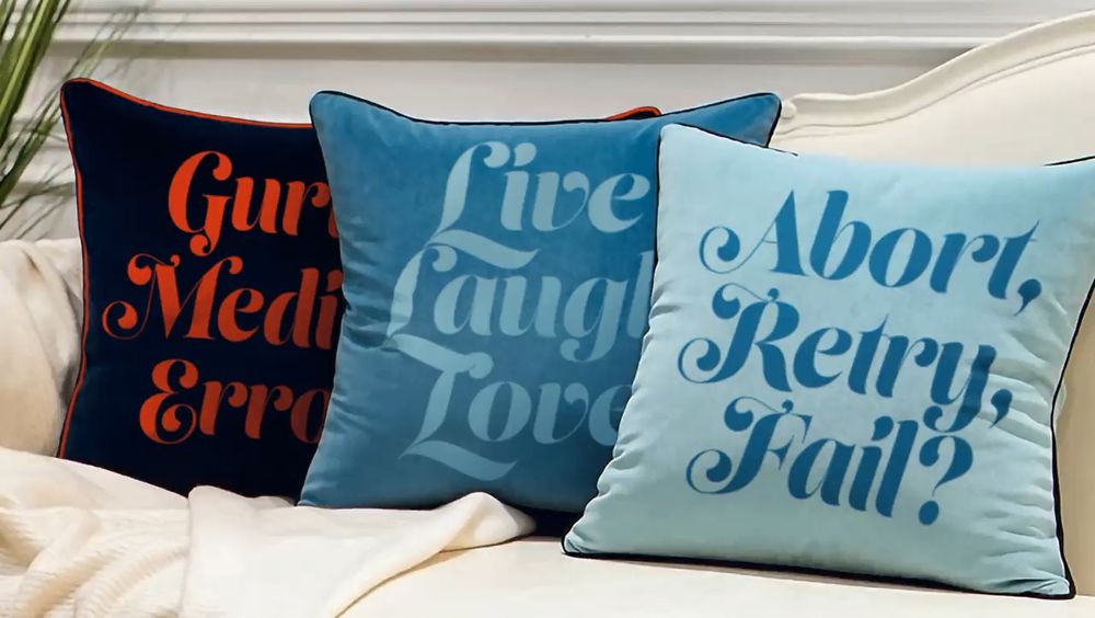 Three pillows with slogans on them. The centremost, LIVE LAUGH LOVE. To the right, ABORT, RETRY, FAIL? and to the left poking out from behind the others, GURU MEDITATION ERROR.