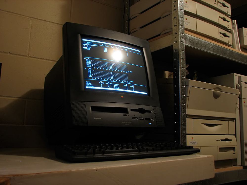 A photo of a black Power Macintosh 5500in a low-lit room, running linux, and showing a shell session with bmon running onscreen. It's a black all-in-one machine with a 15 inch shadow-mask crt, and floppy & CDROM drives on the front, with some volume and brightness controls.