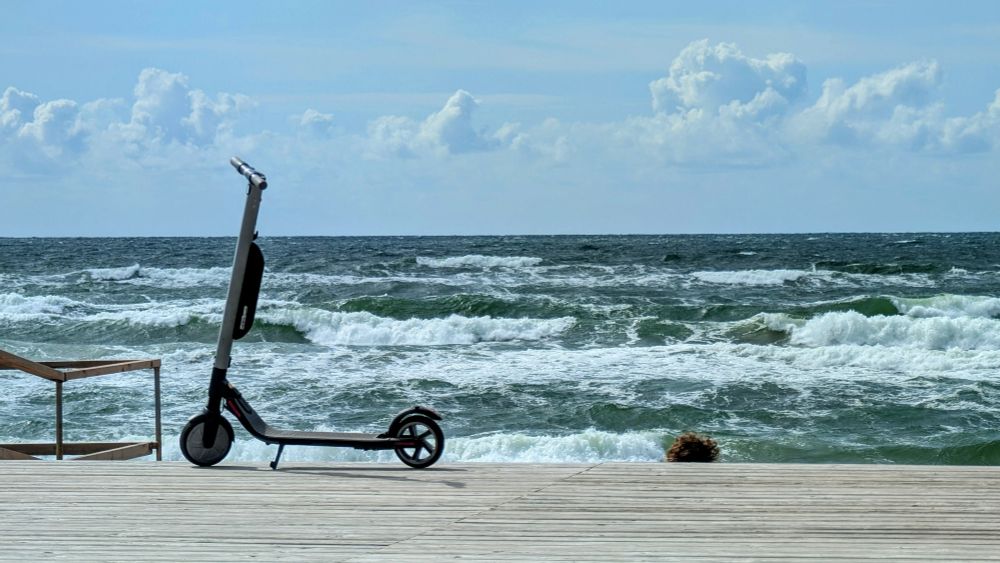 Waves rolling ashore under a hazy blue sky with some fluffy white clouds. In the foreground an electric scooter is parked on a wooden Boardwalk 