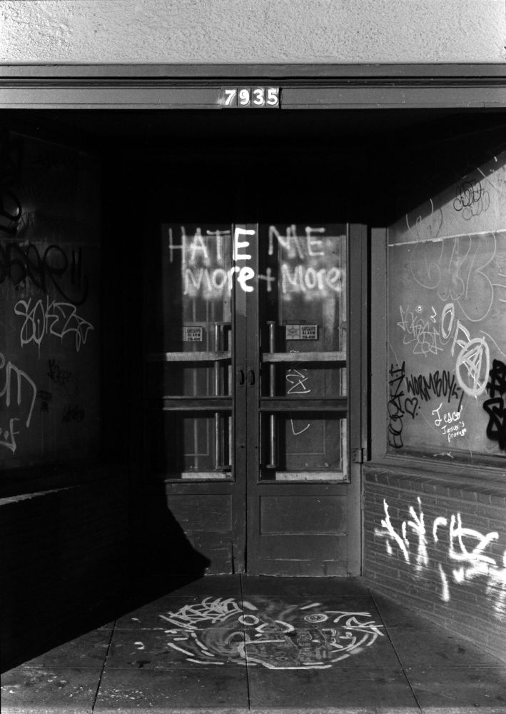 A black and white photograph of a doorway shaded from the left: the front step bears a totemic scrawl of a monstrous face, minor grafitos have left their inscriptions on the brick and glass and contributed their little pieces to the collage texture framing two screen doors shut forever, and, screwed shut tight over locked shop doors of a Reagan vintage and host to the centerpiece of the image, an unaddressed inscription:

“HATE ME
MORE+MORE”

The author of which, in disciplined dispassion or in red hot emotions disregarded the multifaceted topology of their canvas interleaving text with three or more layers of screen and door and glass and blinds.