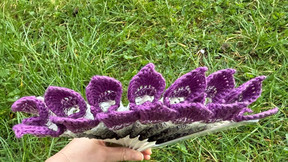Leaf lace crochet hand fan in gradient colours: black - grey - white- purple. Fan is partly closed with green grass in the background. Yarn by crochet uk, pattern by ZomBambie.