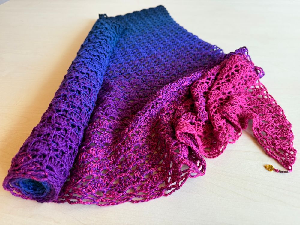 Laid out on a bright table top. A partly rolled up crocheted shawl with gradient colour change: pink - purple - blue - dark blue.