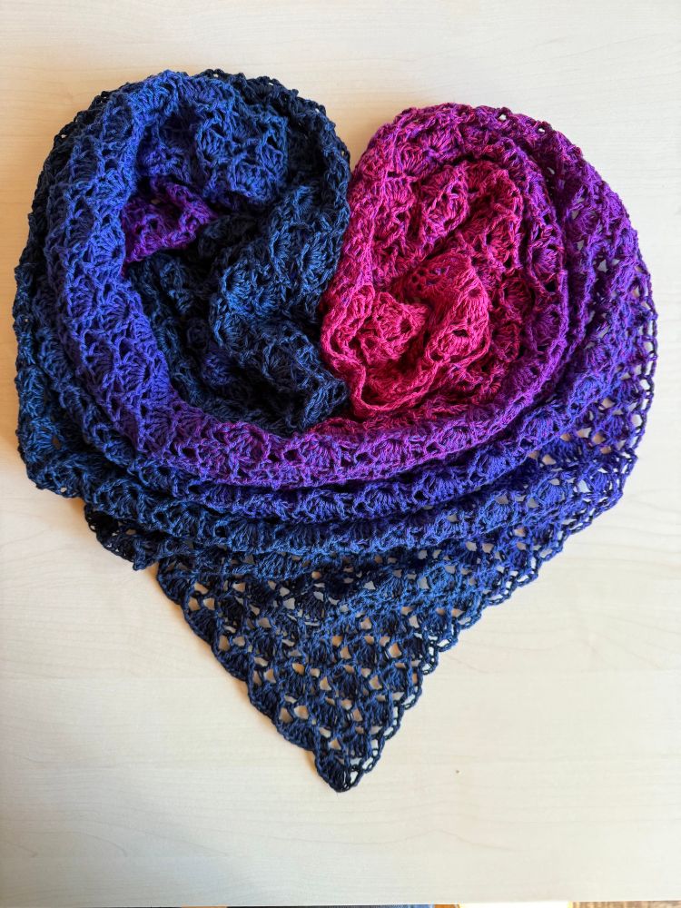 Laid out in heart form on a bright table top. A crocheted shawl with gradient colour change: pink - purple - blue - dark blue.