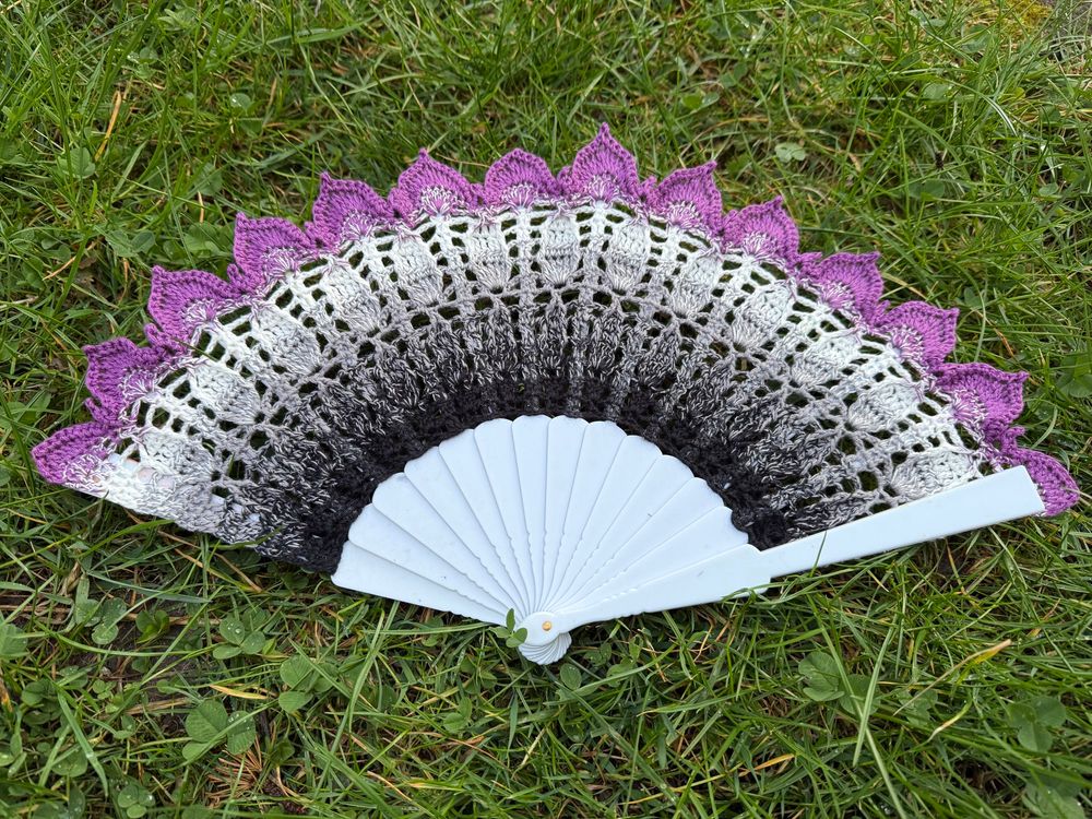 Leaf lace crochet hand fan in gradient colours: black - grey - white- purple. Fan lays on green grass. Yarn by crochet uk, pattern by ZomBambie.