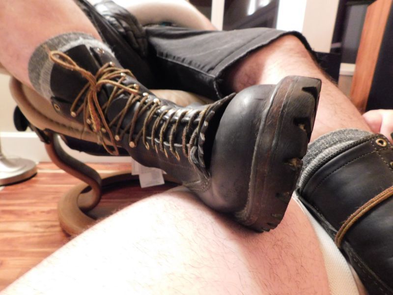 close-up of a Wesco jobmaster tied with wool socks peeking out the top resting on the thighs of a white-skinned person. The Bear in boots is brown with his hairy legs showing with black denim shorts. His right hand is resting on his thigh with a leather glove on you can see up to his belly but not his face. 