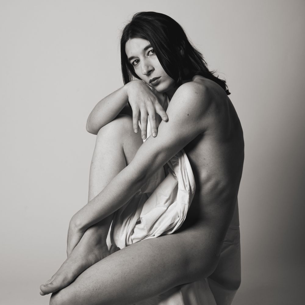 Monochrome photo of a naked trans girl covered in a white sheet while sitting on a chair, studio setting, gray background, she’s looking in camera with a sad face 