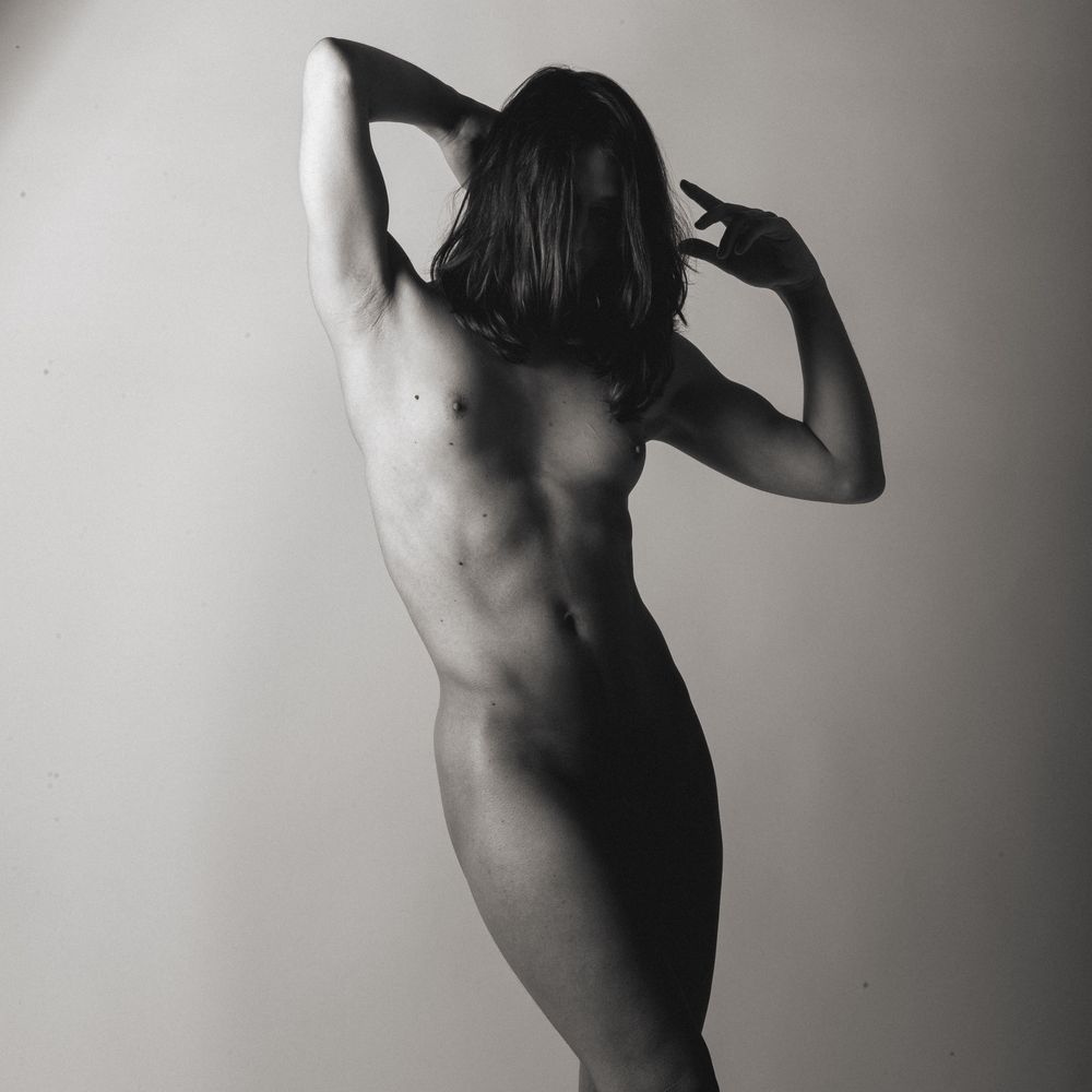 Professional monochrome photo of a naked girl, arms up, face covered by hair, genitals in shadow, posing standing, gray studio background