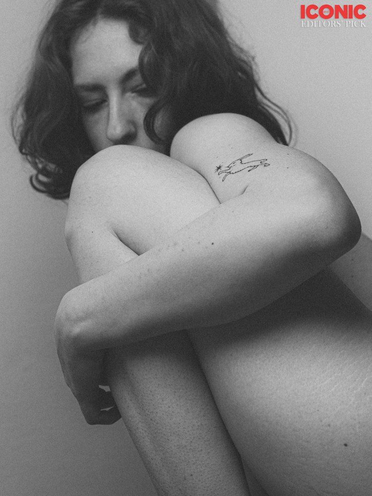 Monochrome professional photo of a naked person hiding behind their knee, showing a slightly tattooed arm and looking down. The model has medium lenght hair and the photo is a medium close up. There’s a red Iconic watermark on the top right