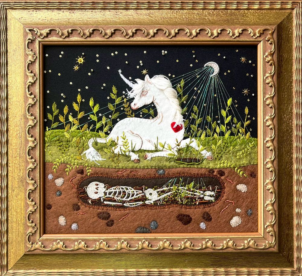 Embroidery of a white unicorn sitting above the grave of a skeleton