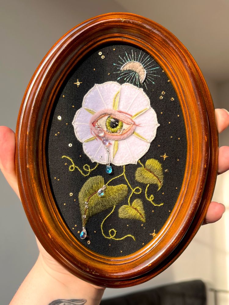 Embroidered white moon flower with green eye in centre, whimsy leaves and dew drop tears coming from the eye using glass beads. Surrounded by stars.