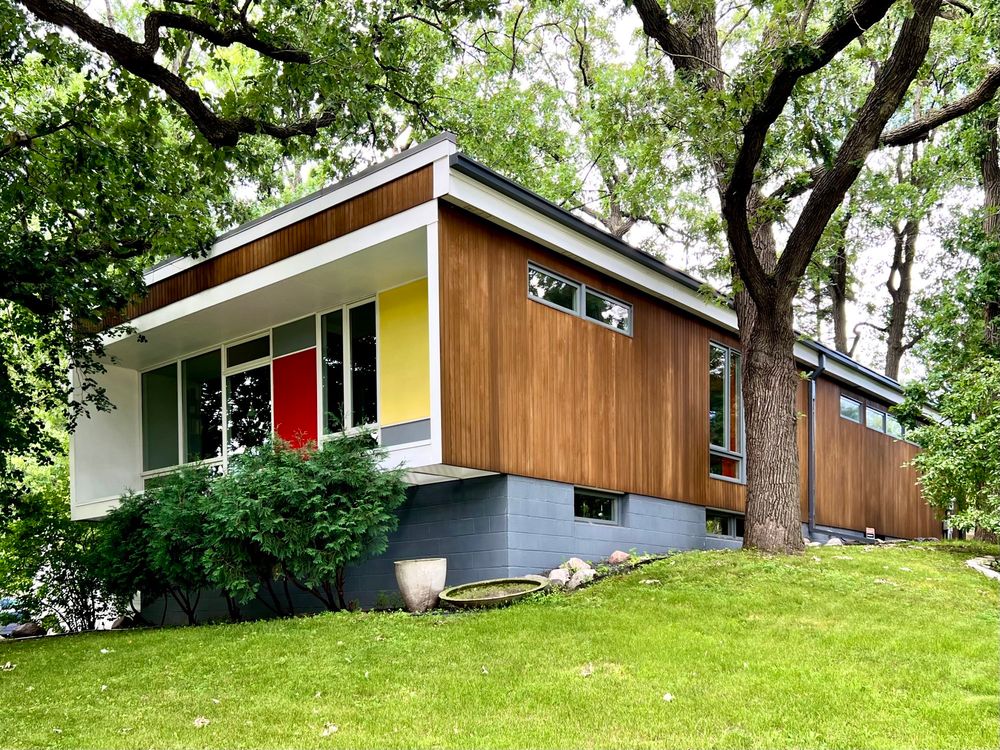 Rectangular, midcentury house with wood siding and a shorter, recessed elevation with multi-colored wall panels.