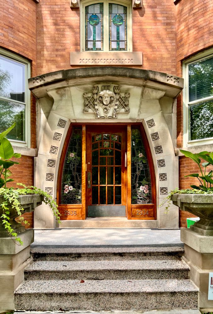 Stone doorway surround that bulges as if being pressed down upon. The top of the doorway features a decorative carving of a woman’s face with flowing hair surrounded by flowers.